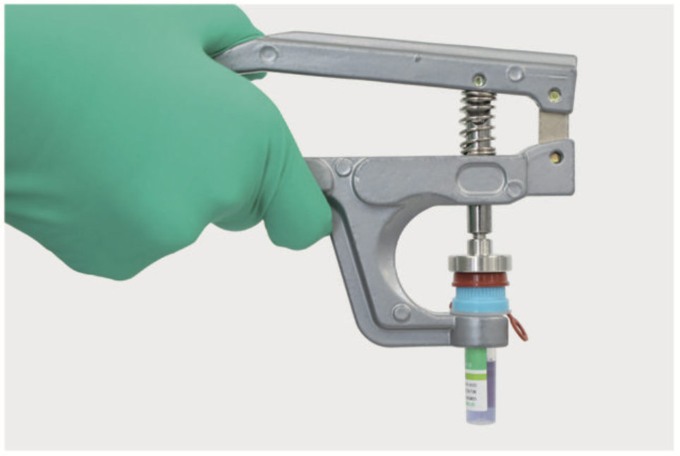 Gloved hand using a spring-loaded medical instrument with a small sample vial attached at the bottom.